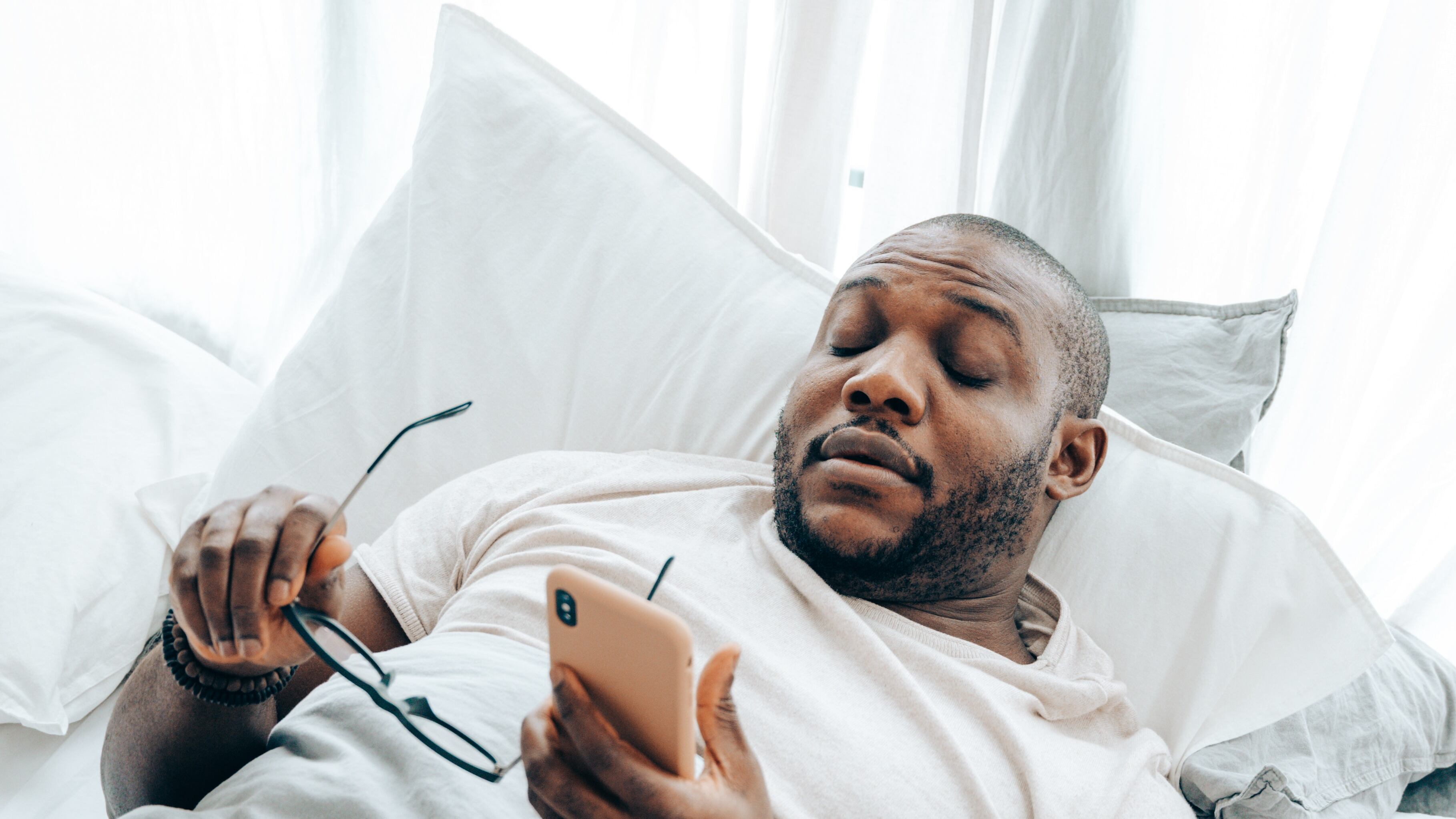 Homem negro deitado segurando óculos com uma mão e celular com outra